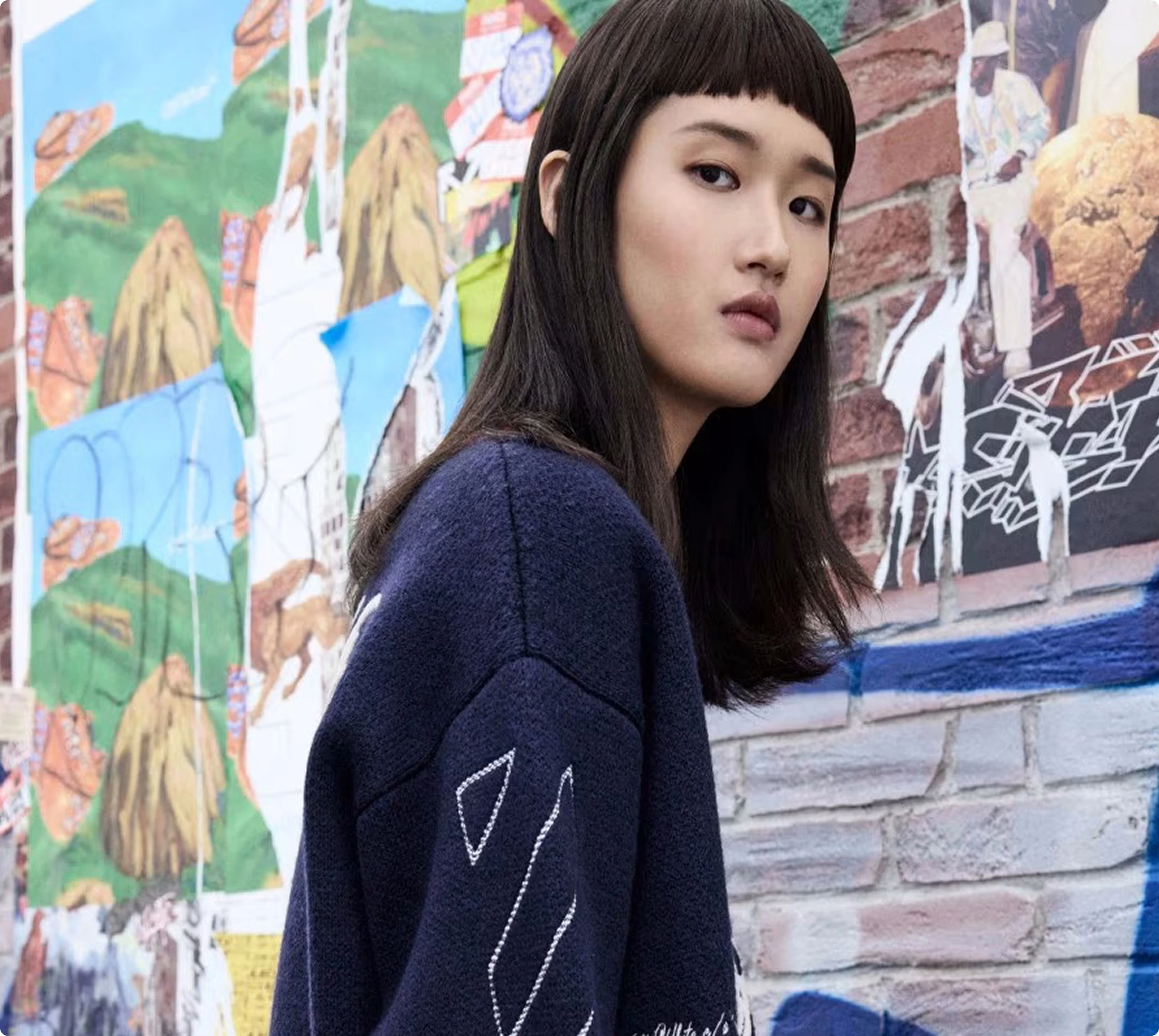 Young woman in navy sweater stands against a vibrant graffiti wall, looking thoughtfully at the camera.