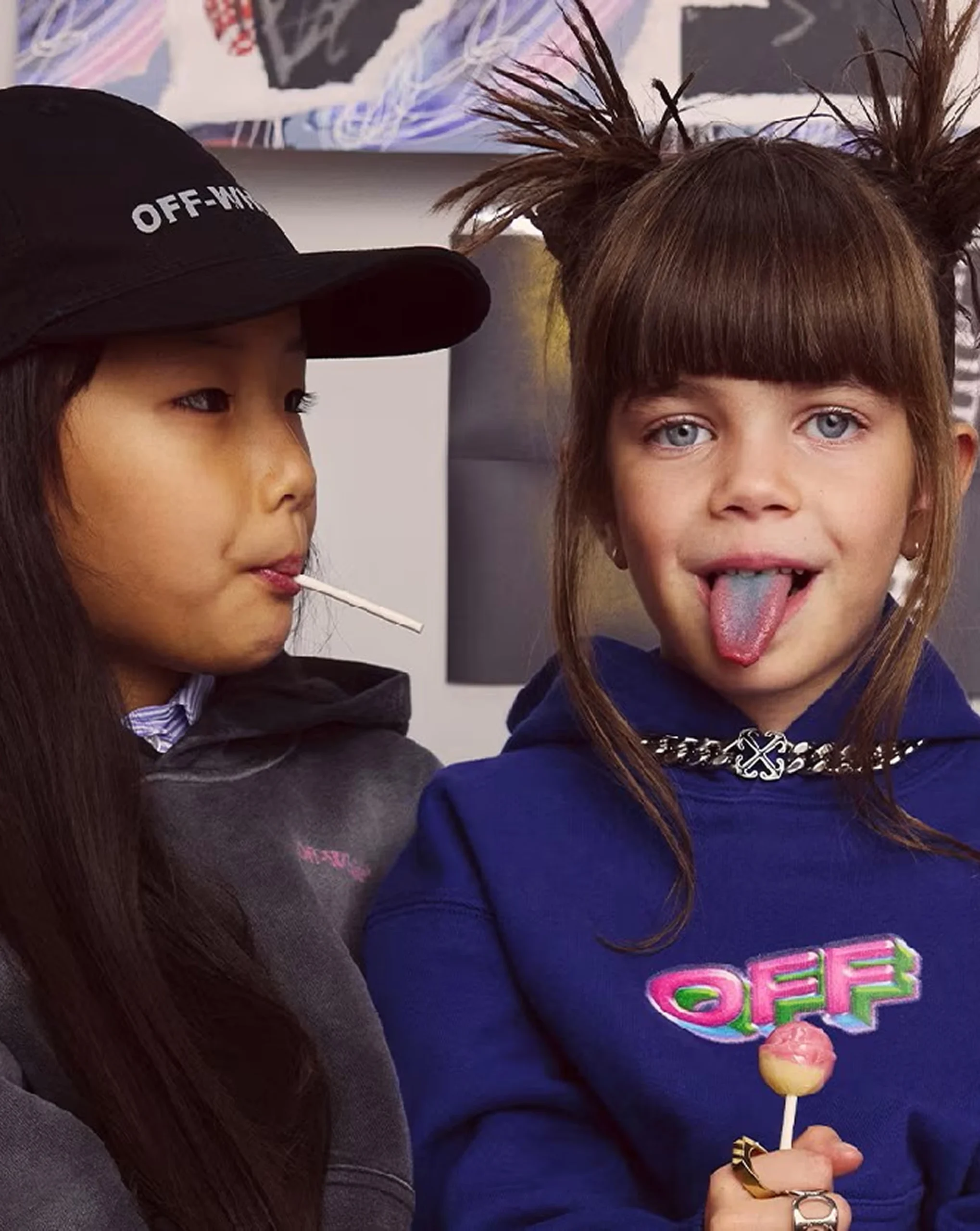 Two kids enjoying lollipops, one with a blue tongue, both wearing stylish streetwear hoodies and caps indoors.