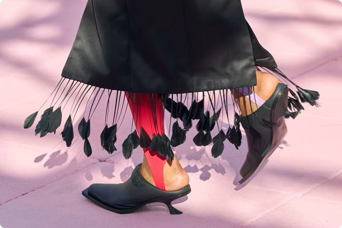 Stylish black dress with feather details, red leggings, and black heeled shoes on pink pavement. Fashion statement ensemble.