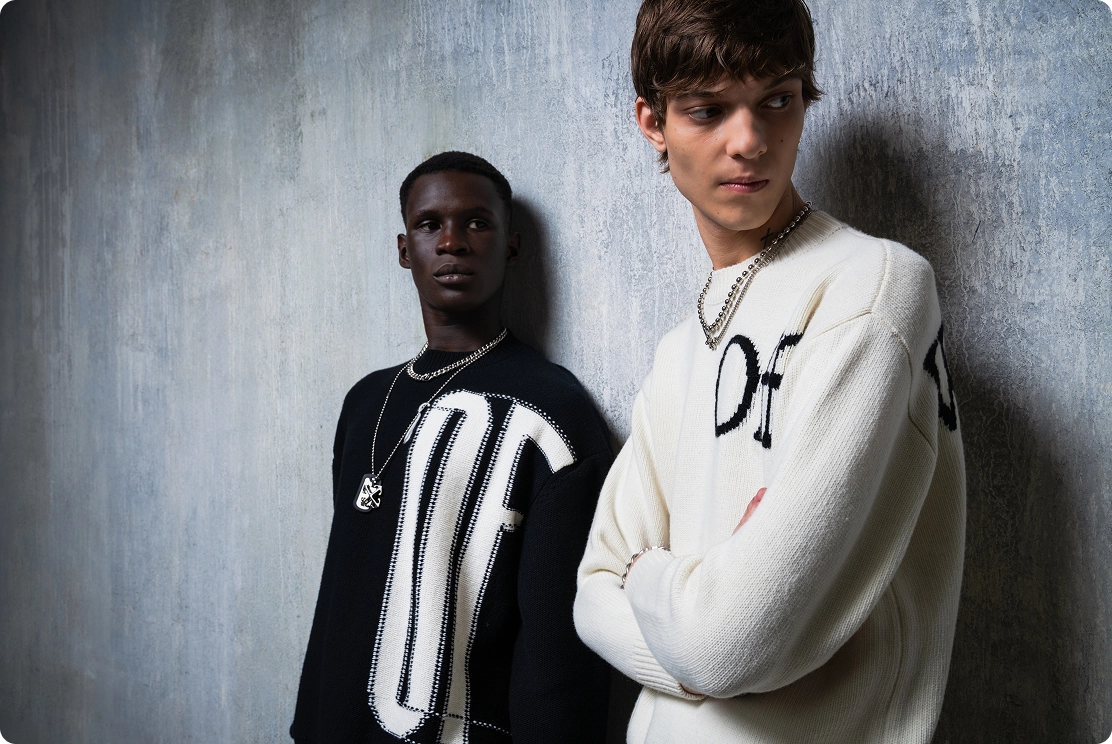 Two models in stylish black and white sweaters pose against a textured wall.