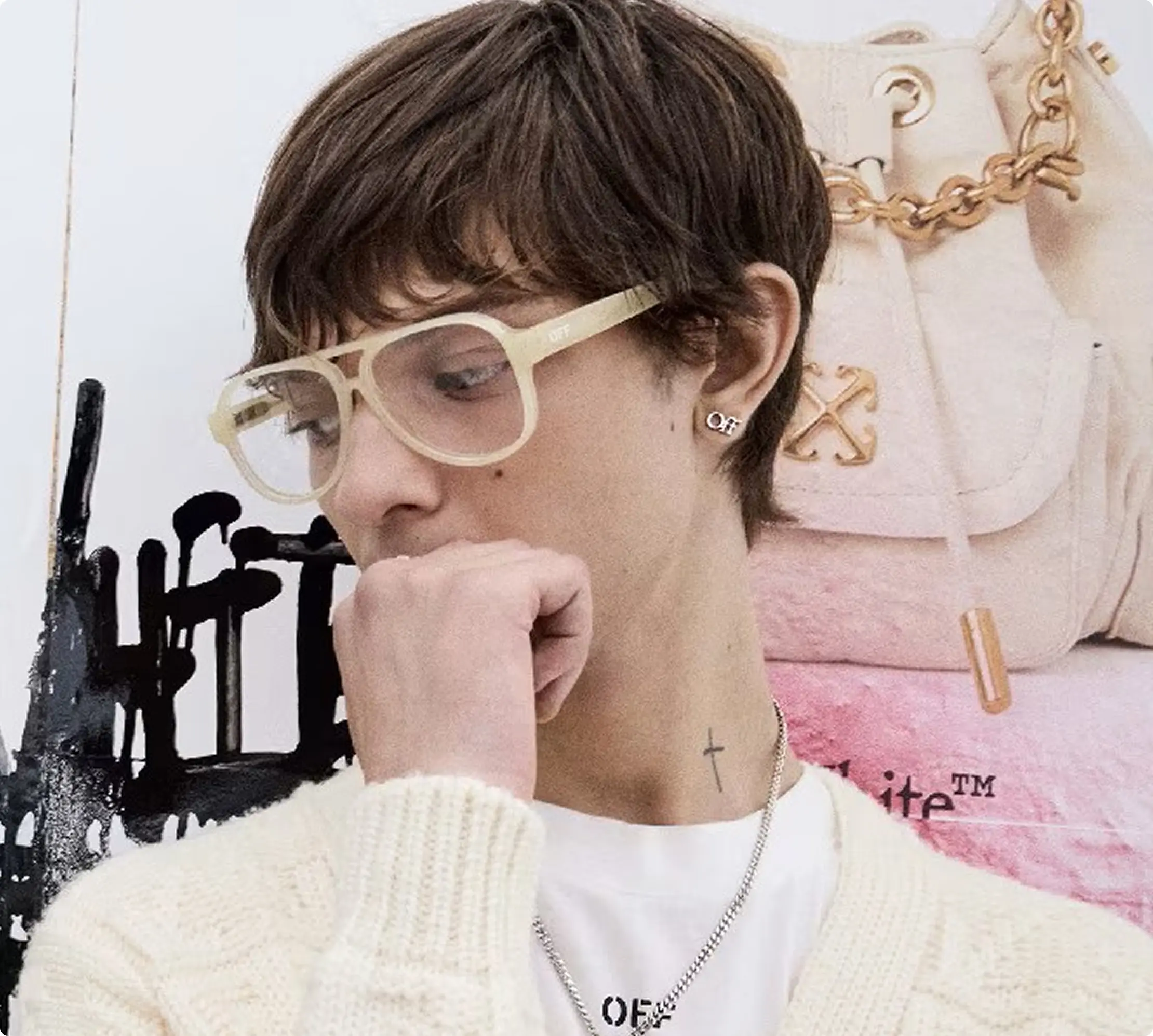 Person wearing clear glasses and a cross necklace, posing thoughtfully by a trendy bag.