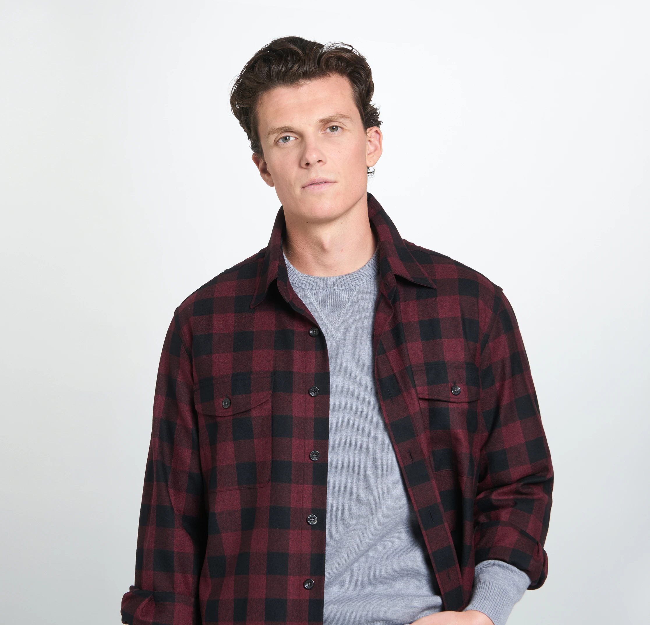 Man in a red and black plaid shirt over a gray sweater, standing against a plain white background.